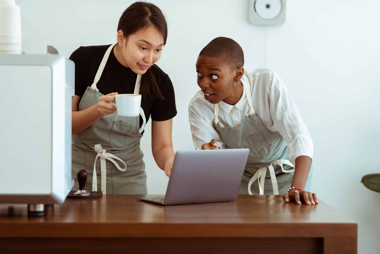 Cheerful surprised diverse colleagues in aprons using modern netbook and sharing business ideas while gathering at table with cup of drink in light cozy kitchen